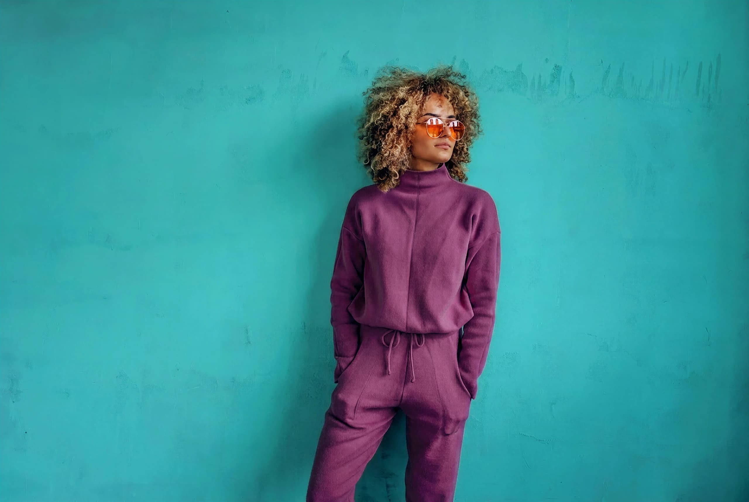 Woman in a purple outfit and sunglasses against a turquoise wall